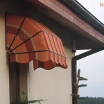 Zdjęcie przedstawia markizę nadokienną o konstrukcji koszowej, zamontowaną na jasnej, tynkowanej elewacji budynku jednorodzinnego bezpośrednio pod okapem dachu. Poszycie markizy wykonane jest z tkaniny w pasy w odcieniach pomarańczu, czerwieni i żółci, co nadaje jej wyrazisty, ciepły wygląd. Konstrukcja opiera się na czarnym, aluminiowym stelażu składającym się z czterech łukowatych ram, które nadają osłonie charakterystyczny, wypukły kształt ćwiartki kuli. Dolna część tkaniny zakończona jest dekoracyjną, swobodnie zwisającą falbaną o falistym brzegu, obszytą ciemną lamówką pasującą do koloru stelaża. Markiza jest zamontowana nad wąskim oknem, a w jej sąsiedztwie widoczny jest fragment brązowej rynny spustowej oraz ciemna podbitka dachowa. Tło stanowi zamglony krajobraz z linią lasu, co sugeruje lokalizację budynku na wzniesieniu lub w otwartej przestrzeni.