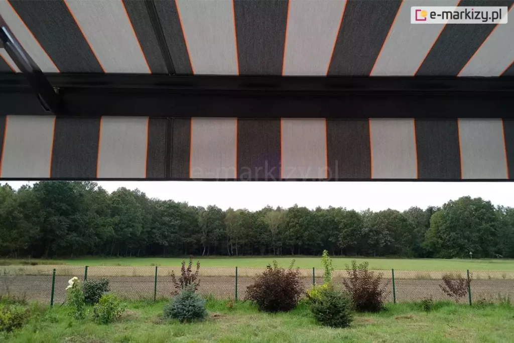 widok z wnętrza tarasu na otaczający krajobraz, częściowo przesłonięty przez nowoczesną markizę tarasową Palladio Aluprof. Poszycie markizy wykonane z tkaniny akrylowej powlekanej teflonem, charakteryzuje się rytmicznym układem szerokich pasów w kolorze ciemnografitowym oraz jasnoszarym, które są oddzielone od siebie cienkimi, pionowymi liniami w kolorze intensywnej pomarańczy. Materiał jest napięty na czarnej, aluminiowej konstrukcji, której poprzeczna belka czołowa widoczna jest w górnej części kadru. Markiza posiada również prostą falbanę o tym samym wzorze, która zwisa z profilu, ograniczając dostęp światła słonecznego padającego pod niskim kątem. W tle, pod linią osłony, rozpościera się widok na zielony ogród z niskimi nasadzeniami krzewów ozdobnych o bordowych i zielonych liściach oraz iglaków. Za niskim ogrodzeniem z siatki widać pas zaoranego pola, jasną łąkę oraz gęstą ścianę liściastego lasu pod zachmurzonym niebem. Perspektywa zdjęcia podkreśla, jak zastosowanie markizy o nowoczesnym designie pozwala na stworzenie komfortowej, zacienionej przestrzeni wypoczynkowej z zachowaniem pełnego kontaktu z naturą i widokiem na otwartą przestrzeń.