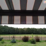 widok z wnętrza tarasu na otaczający krajobraz, częściowo przesłonięty przez nowoczesną markizę tarasową Palladio Aluprof. Poszycie markizy wykonane z tkaniny akrylowej powlekanej teflonem, charakteryzuje się rytmicznym układem szerokich pasów w kolorze ciemnografitowym oraz jasnoszarym, które są oddzielone od siebie cienkimi, pionowymi liniami w kolorze intensywnej pomarańczy. Materiał jest napięty na czarnej, aluminiowej konstrukcji, której poprzeczna belka czołowa widoczna jest w górnej części kadru. Markiza posiada również prostą falbanę o tym samym wzorze, która zwisa z profilu, ograniczając dostęp światła słonecznego padającego pod niskim kątem. W tle, pod linią osłony, rozpościera się widok na zielony ogród z niskimi nasadzeniami krzewów ozdobnych o bordowych i zielonych liściach oraz iglaków. Za niskim ogrodzeniem z siatki widać pas zaoranego pola, jasną łąkę oraz gęstą ścianę liściastego lasu pod zachmurzonym niebem. Perspektywa zdjęcia podkreśla, jak zastosowanie markizy o nowoczesnym designie pozwala na stworzenie komfortowej, zacienionej przestrzeni wypoczynkowej z zachowaniem pełnego kontaktu z naturą i widokiem na otwartą przestrzeń.