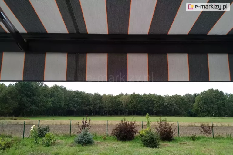 widok z wnętrza tarasu na otaczający krajobraz, częściowo przesłonięty przez nowoczesną markizę tarasową Palladio Aluprof. Poszycie markizy wykonane z tkaniny akrylowej powlekanej teflonem, charakteryzuje się rytmicznym układem szerokich pasów w kolorze ciemnografitowym oraz jasnoszarym, które są oddzielone od siebie cienkimi, pionowymi liniami w kolorze intensywnej pomarańczy. Materiał jest napięty na czarnej, aluminiowej konstrukcji, której poprzeczna belka czołowa widoczna jest w górnej części kadru. Markiza posiada również prostą falbanę o tym samym wzorze, która zwisa z profilu, ograniczając dostęp światła słonecznego padającego pod niskim kątem. W tle, pod linią osłony, rozpościera się widok na zielony ogród z niskimi nasadzeniami krzewów ozdobnych o bordowych i zielonych liściach oraz iglaków. Za niskim ogrodzeniem z siatki widać pas zaoranego pola, jasną łąkę oraz gęstą ścianę liściastego lasu pod zachmurzonym niebem. Perspektywa zdjęcia podkreśla, jak zastosowanie markizy o nowoczesnym designie pozwala na stworzenie komfortowej, zacienionej przestrzeni wypoczynkowej z zachowaniem pełnego kontaktu z naturą i widokiem na otwartą przestrzeń.