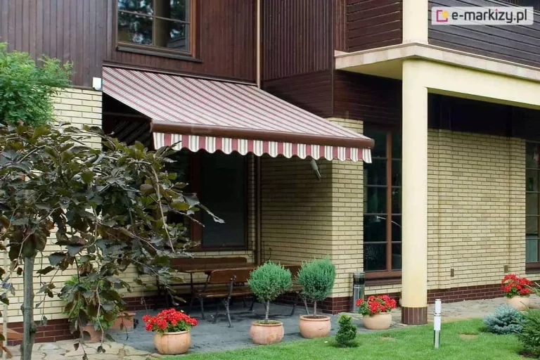 Zdjęcie przedstawia strefę wypoczynkową na tarasie przylegającym do budynku o mieszanej elewacji, wykonanej z jasnej cegły oraz ciemnobrązowych paneli drewnianych. Nad częścią tarasu zamontowana jest klasyczna, w pełni rozwinięta markiza tarasowa Palladio, charakteryzująca się poszyciem w pionowe pasy w kolorach bordowym, białym i beżowym. Dolna krawędź tkaniny wykończona jest dekoracyjną, falistą falbaną, która nawiązuje do kolorystyki głównego materiału. Konstrukcja markizy, w tym belka przednia, utrzymana jest w kolorze brązowym, pasującym do stolarki okiennej i drewnianych elementów elewacji. Pod markizą znajduje się drewniany stół z ławami, ustawiony na podłożu z szarej kostki lub płyt kamiennych. Przestrzeń wokół tarasu jest bogato zdobiona roślinnością: w glinianych donicach rosną jaskrawoczerwone pelargonie oraz formowane w kule niskie krzewy iglaste. Po lewej stronie kadru widoczne jest ozdobne drzewko o ciemnych, bordowych liściach, a na pierwszym planie znajduje się starannie utrzymany zielony trawnik. Całość kompozycji ukazuje przytulny i funkcjonalny kącik do odpoczynku na świeżym powietrzu w słoneczny dzień.