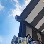 Widok belki przedniej markizy Palladio w kolorze brązowym z poszyciem i falowaną falbaną w pasy kremowe i brązowe na tle kamienic i pięknego, pogodnego błękitnego nieba z kilkoma białymi obłokami.