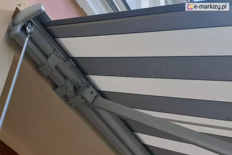 Zbliżenie lewej strony markizy Palladio Aluprof z grafitową konstrukcją oraz rozwiniętym poszyciem w pasy o trzech odcieniach szarości zamocowanej na elewacji nad wnęką okienną. Od boku widoczna zaślepka boczna z PCV oraz zaczep oczkowy przekładni korbowej do sterowania ręcznego, z zaczepioną o nią korbą. W głębi widoczne także, mocowania kasety, uchwyt ścienny oraz mocowanie ramienia markizy do belki kwadratowej.