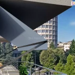 markiza Italia bez kasety Aluprof z antracytową konstrukcją oraz ramionami mocowanymi do ściany zawieszona nad wyjściem na taras, pod płytą balkonu wyższego piętra budynku. ramiona opuszczone pod katem około 50 stopni napinają szara tkaninę poszycia, która osłania wyjście na taras. W tle za tarasem widoczny balkon sąsiedniego mieszkania, ulica o średnim natężeniu ruchu częściowo przesłonięta licznymi drzewami oraz sąsiednie budynki, w tym widoczny na tle markizy wysoki obiekt o zaokrąglonej fasadzie.