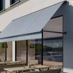 Wizualizacja zewnętrznego ogródka restauracyjnego lub kawiarnianego usytuowanego bezpośrednio przy nowoczesnym budynku o jasnej, tynkowanej elewacji. Nad dużymi witrynami lokalu zamontowana jest szeroka markiza fasadowa w kolorze jasnoszarym. Jest to model markizy z ramionami opadowymi, których punkty montażowe znajdują się po bokach okien, co pozwala na stabilne nachylenie poszycia pod kątem. Markiza jest w pełni rozwinięta, osłaniając wnętrze lokalu oraz część stolików przed słońcem. W witrynach odbija się ulica, biały samochód osobowy oraz zieleń miejska. Przed lokalem, na chodniku, ustawiono rzędy drewnianych stolików i ław o prostej konstrukcji z metalowymi stelażami. Na stolikach widoczne są drobne elementy dekoracyjne, takie jak małe doniczki z sukulentami oraz karafki z oliwą. Podłoże stanowi jasna nawierzchnia brukowa. Budynek charakteryzuje się regularnym rytmem okien na wyższych kondygnacjach, podkreślonych ciemniejszymi pasami elewacji. Oświetlenie wskazuje na słoneczne popołudnie, a cienie rzucane przez markizę i meble są wyraźne i krótkie.