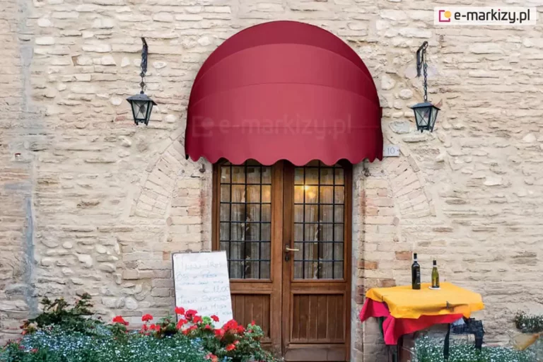 Zdjęcie przedstawia wejście do klimatycznego lokalu, prawdopodobnie restauracji lub winiarni, usytuowane w starej, kamienno-ceglanej ścianie budynku o rustykalnym charakterze. Nad dwuskrzydłowymi, drewnianymi drzwiami z licznymi przeszkleniami i ciemnymi szprosami zamontowana jest klasyczna markiza koszowa o półkolistym kształcie. Tkanina markizy ma intensywny, bordowy lub ciemnoczerwony kolor, a jej dolna krawędź jest wykończona ozdobną, pofalowaną falbaną. Elewacja wokół drzwi wykonana jest z jasnych, nieregularnych kamieni i cegieł, tworząc nad wejściem łukową konstrukcję. Po obu stronach markizy, na ścianie, zamocowane są stylizowane, czarne latarnie o klasycznym kształcie, które pełnią funkcję oświetlenia zewnętrznego. Po prawej stronie drzwi znajduje się mały stolik nakryty żółtym obrusem z czerwoną podkładką, na którym stoją dwie butelki wina. Po lewej stronie widoczna jest biała tablica z ręcznie wypisanym menu oraz fragment kwietnika z czerwonymi pelargoniami i drobnymi, niebieskimi kwiatami. Całość kompozycji emanuje śródziemnomorskim, przytulnym nastrojem, podkreślonym przez ciepłe światło przebijające przez szyby drzwi.