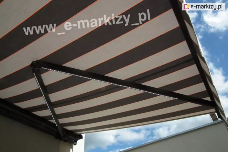 Zdjęcie prezentuje zbliżenie na detale konstrukcyjne od spodu, jakie posiada markiza rozsuwana Palladio, zamontowana na białej elewacji budynku. Poszycie markizy wykonane jest z tkaniny w szerokie, poziome pasy w kolorze ciemnobrązowym i beżowym, które są oddzielone od siebie cienkimi, kontrastującymi liniami w kolorze intensywnej pomarańczy. Najbardziej charakterystycznym elementem widocznym na fotografii są solidne ramiona przegubowe wykonane z aluminium lakierowanego proszkowo na głęboki kolor antracytowy. Wzdłuż profili ramion zamocowano nowoczesne oświetlenie LED w formie taśm, które emitują jasne, punktowe światło skierowane w dół, co pozwala na komfortowe korzystanie z tarasu po zmroku. Konstrukcja ramion jest masywna i technicznie zaawansowana, z widocznymi przegubami i mechanizmem naprężającym tkaninę. W dolnej części kadru widoczny jest fragment krawędzi dachu sąsiedniego obiektu oraz jasnobłękitne niebo z rozproszonymi, białymi chmurami. Całość ujęcia podkreśla wysoką jakość wykończenia oraz funkcjonalność dodatkowego oświetlenia w systemie Palladio.