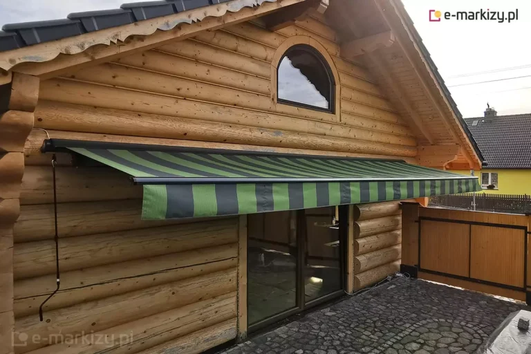 Zdjęcie prezentuje budynek wykonany z grubych, naturalnych bali drewnianych, na którego elewacji zamontowano szeroką markizę Adagio. Markiza jest częściowo rozwinięta, a jej poszycie charakteryzuje się wzorem w pionowe pasy w dwóch odcieniach zieleni oraz czerni. Konstrukcja markizy Adagio, w tym kaseta ochronna oraz profil frontowy, jest w kolorze ciemnego grafitu. Z lewej strony profilu frontowego zwiesza się długa, metalowa korba służąca do ręcznego rozkładania i składania zadaszenia. Poniżej tkaniny widoczna jest falbana o tym samym wzorze co główny materiał. Markiza Adagio osłania duże, przeszkolone drzwi tarasowe w ciemnej ramie. Przed budynkiem znajduje się taras wyłożony ciemną kostką granitową o nieregularnym kształcie. Powyżej markizy, w szczycie drewnianej ściany, umieszczone jest półokrągłe okno, a dach budynku pokryty jest ciemną dachówką ceramiczną z ozdobnie wykończonymi deskami czołowymi. W tle, po prawej stronie, widać fragment drewnianego ogrodzenia oraz sąsiedni dom z żółtą elewacją pod pochmurnym niebem.