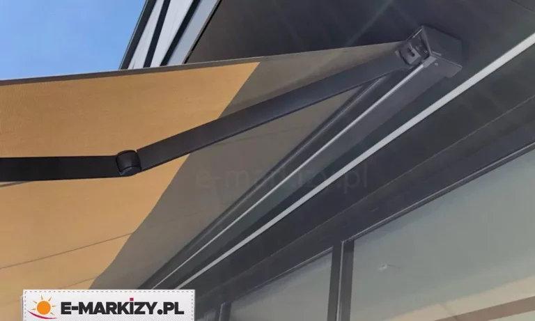 Zdjęcie prezentuje nowoczesną markizę ścienną zainstalowaną na ciemnej elewacji budynku, przedstawioną z perspektywy od dołu, co eksponuje jej mechanizm i ramię przegubowe. Markiza Moderno posiada konstrukcję w kolorze antracytowym o matowym wykończeniu, a jej ramię jest w pełni wyprostowane, podtrzymując napięte, beżowe poszycie o wyraźnym splocie tkaniny. Materiał markizy rzuca geometryczny cień na ciemną ścianę oraz dużą szklaną witrynę znajdującą się poniżej zadaszenia. System mocowania ramienia do profilu nośnego charakteryzuje się minimalistyczną, kanciastą formą, typową dla współczesnej architektury. W lewym górnym rogu kadru widoczny jest fragment czystego, błękitnego nieba, co podkreśla funkcję ochronną produktu w słoneczny dzień.
