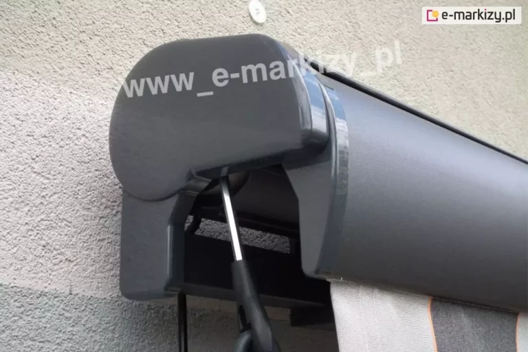 Lewy bok markizy Palladio w kolorze srebrzysty grafit z zaślepką boczną z pcv oraz wystającą z zamkniętej belki przedniej falbaną w pasy o dwóch odcieniach szarości. Pod zaślepką widoczny przegub przekładni korbowej, do którego zaczepia się korbę napędu ręcznego.