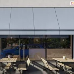 Wizualizacja przedstawiająca nowoczesną markizę sklepową z ramionami opadowymi, zamontowaną nad witryną lokalu usługowego lub restauracji. Markiza posiada gładkie poszycie w kolorze jasnoszarym, które jest w pełni rozwinięte i nachylone pod stałym kątem, zapewniając ochronę przeciwsłoneczną. Konstrukcja wsporcza, obejmująca ramiona boczne oraz belkę frontową, wykonana jest z profili aluminiowych w kolorze czarnym lub bardzo ciemnym grafitowym. Ramiona są przymocowane do jasnej, tynkowanej elewacji budynku po obu stronach dużego przeszklenia. W witrynie odbija się fragment ulicy oraz zaparkowany przy krawężniku niebieski samochód typu SUV. Przed lokalem, na brukowanym podłożu, ustawiono drewniane stoliki kawiarniane z dopasowanymi ławami o metalowych, ozdobnych stelażach. Na stolikach znajdują się drobne akcesoria, takie jak białe doniczki z roślinami. Scena ukazuje słoneczny dzień, co podkreślają wyraźne, ukośne cienie rzucane przez meble oraz krawędź markizy na elewację i chodnik.