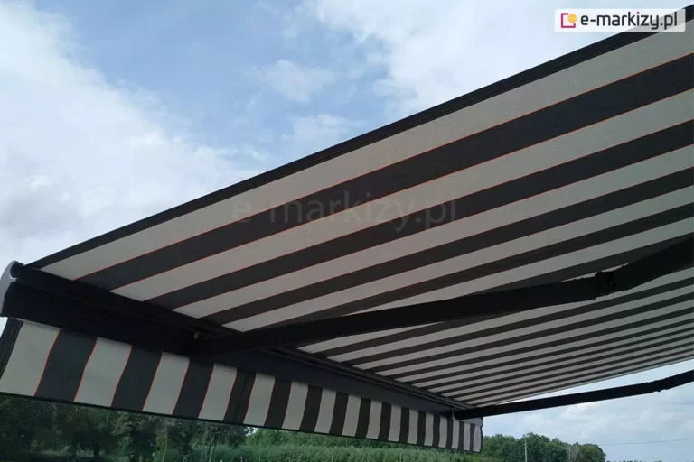 Zdjęcie prezentuje zbliżenie na spód rozłożonej markizy tarasowej Palladio, ukazujące detale jej poszycia oraz konstrukcji wsporczej na tle zachmurzonego nieba. Akrylowa tkanina markizy ozdobiona jest wzorem w szerokie, poziome pasy w kolorach ciemnoszarym i jasnoszarym, które są oddzielone od siebie cienkimi, kontrastującymi prążkami w kolorze pomarańczowym. Markiza posiada prostą, krótką falbanę, która powtarza ten sam pasiasty wzór i kolorystykę. Konstrukcja nośna widoczna pod materiałem składa się z solidnych, czarnych ramion przegubowych o matowym wykończeniu, które stabilizują napięcie tkaniny. W dolnej części kadru, poniżej krawędzi markizy, widoczna jest linia horyzontu z gęstym pasem zielonych drzew. Perspektywa zdjęcia z dołu podkreśla dużą powierzchnię zadaszenia oraz techniczną precyzję mechanizmu ramion.