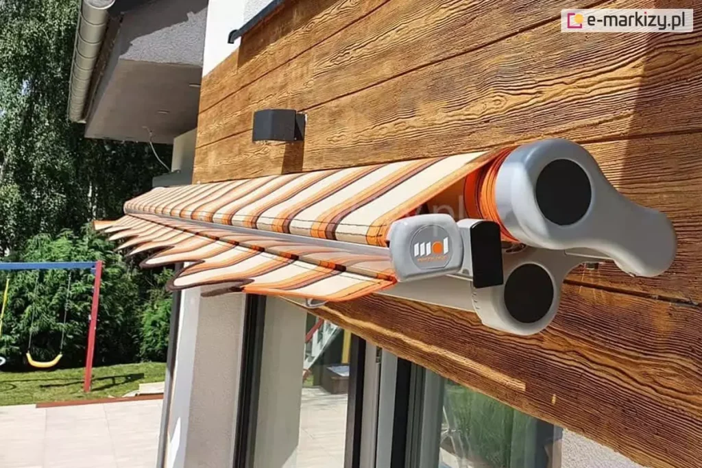 Zdjęcie prezentuje perspektywę boczną na markizę tarasową Allegro, w stanie niemal całkowitego zwinięcia. Widok z bliska pozwala na analizę technicznych komponentów systemu bezkasetowego, zamontowanego na elewacji wykończonej deską kompozytową o wyraźnej strukturze drewna w odcieniu miodowego brązu. Na pierwszym planie widoczna jest masywna, boczna głowica konstrukcji wykonana z odlewu aluminium w kolorze jasnoszarym, o zaokrąglonym kształcie z czarnymi maskownicami przegubów. Tuż obok znajduje się profil czołowy, na którym zamocowano dekoracyjną, falowaną falbanę z ciemnobrązową lamówką. Tkanina markizy, zwinięta na rurze nawojowej, charakteryzuje się wzorem w pionowe pasy w kolorystyce pomarańczowej, beżowej i ciemnobrązowej. Powyżej markizy na ścianie zainstalowano nowoczesną, czarną lampę zewnętrzną o geometrycznym profilu. W tle, po lewej stronie kadru, widoczny jest fragment ogrodu z intensywnie zielonymi drzewami i krzewami oraz fragment konstrukcji dziecięcej huśtawki, co podkreśla domowy, rekreacyjny charakter otoczenia.