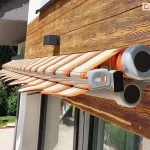 Zdjęcie prezentuje perspektywę boczną na markizę tarasową Allegro, w stanie niemal całkowitego zwinięcia. Widok z bliska pozwala na analizę technicznych komponentów systemu bezkasetowego, zamontowanego na elewacji wykończonej deską kompozytową o wyraźnej strukturze drewna w odcieniu miodowego brązu. Na pierwszym planie widoczna jest masywna, boczna głowica konstrukcji wykonana z odlewu aluminium w kolorze jasnoszarym, o zaokrąglonym kształcie z czarnymi maskownicami przegubów. Tuż obok znajduje się profil czołowy, na którym zamocowano dekoracyjną, falowaną falbanę z ciemnobrązową lamówką. Tkanina markizy, zwinięta na rurze nawojowej, charakteryzuje się wzorem w pionowe pasy w kolorystyce pomarańczowej, beżowej i ciemnobrązowej. Powyżej markizy na ścianie zainstalowano nowoczesną, czarną lampę zewnętrzną o geometrycznym profilu. W tle, po lewej stronie kadru, widoczny jest fragment ogrodu z intensywnie zielonymi drzewami i krzewami oraz fragment konstrukcji dziecięcej huśtawki, co podkreśla domowy, rekreacyjny charakter otoczenia.
