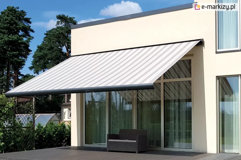 Zdjęcie prezentuje nowoczesną markizę tarasową Palladio Aluprof zainstalowaną na elewacji piętrowego budynku o minimalistycznej architekturze. Markiza posiada duży wysięg i jest pokryta tkaniną w szerokie, pionowe pasy w odcieniach bieli i jasnej szarości. Konstrukcja markizy, w tym kaseta i przednia belka, wykonana jest z metalu w kolorze antracytowym, co kontrastuje z jasną, kremową elewacją domu. Duży kąt nachylenia markizy zapewnia pełne zacienienie wyjścia na taras umieszczone w dużym przeszkleniu na fasadzie. Pod markizą znajduje się taras wykonany z ciemnobrązowych desek kompozytowych, na którym ustawiono pojedynczą, dwuosobową sofę z ciemnej plecionki z jasnym siedziskiem. Tło dla mebla stanowią duże, przeszklone drzwi tarasowe i okna, w których odbija się zieleń ogrodu. Po lewej stronie widoczne jest gęste ogrodzenie z siatki porośnięte roślinnością oraz wysokie sosny na tle błękitnego, lekko zachmurzonego nieba. Architektura budynku charakteryzuje się prostymi liniami i brakiem zbędnych zdobień, co podkreśla funkcjonalny i luksusowy charakter prezentowanej osłony przeciwsłonecznej.
