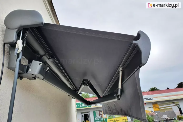 Zawieszona na ścianie budynku z tynkiem strukturalnym w kolorze białym markiza Palladio Aluprof z grafitową konstrukcją oraz tkaniną i prostą falbaną w kolorze Antracita z widocznymi przy niewielkim rozsunięciu skrzyżowanymi ramionami, które składają się przy belce jedno nad drugim. Po lewej widoczny zaczep oczkowy z korbą do ręcznego sterowania.