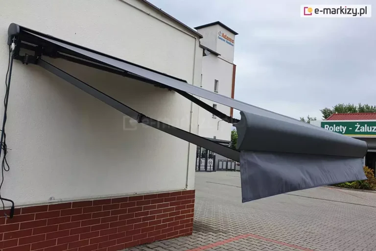 Grafitowa markiza Palladio Aluprof zamocowana na ścianie budynku z ceglaną podmurówką, markiza z ramionami krzyżowymi, z których lewy wysięgnik zainstalowany jest niżej niż prawy, dzięki czemu składają się one równolegle.