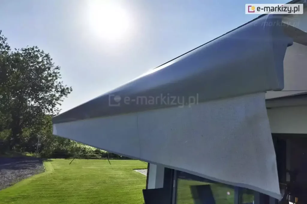 Zbliżenie na belkę przednią markizy tarasowej Palladio zamontowanej nad oknem budynku. Konstrukcja wykonana jest z aluminium w kolorze srebrnym, a po prawej stronie belkę zamyka zaślepka z PCV w charakterystycznym kształcie. Z przedniego profilu markizy wysunięta jest pionowa falbana wykonana z tkaniny akrylowej w kolorze jasnoszarym, która pozwala na częściową widoczność przy jednoczesnym rozpraszaniu promieni słonecznych. W tle widoczny jest fragment zadbanego ogrodu z zielonym trawnikiem oraz gęstymi drzewami liściastymi pod czystym, błękitnym niebem w pełnym słońcu. Światło słoneczne odbija się od górnej powierzchni kasety, podkreślając jej metaliczną strukturę. Po prawej stronie zdjęcia widoczny jest fragment ramy okiennej oraz ciemniejsze wnętrze pomieszczenia.