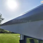 Zbliżenie na belkę przednią markizy tarasowej Palladio zamontowanej nad oknem budynku. Konstrukcja wykonana jest z aluminium w kolorze srebrnym, a po prawej stronie belkę zamyka zaślepka z PCV w charakterystycznym kształcie. Z przedniego profilu markizy wysunięta jest pionowa falbana wykonana z tkaniny akrylowej w kolorze jasnoszarym, która pozwala na częściową widoczność przy jednoczesnym rozpraszaniu promieni słonecznych. W tle widoczny jest fragment zadbanego ogrodu z zielonym trawnikiem oraz gęstymi drzewami liściastymi pod czystym, błękitnym niebem w pełnym słońcu. Światło słoneczne odbija się od górnej powierzchni kasety, podkreślając jej metaliczną strukturę. Po prawej stronie zdjęcia widoczny jest fragment ramy okiennej oraz ciemniejsze wnętrze pomieszczenia.