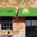 Dwie trójkątne markizy koszowe z poszyciem i prostą falbaną wykonaną z zielonej tkaniny akrylowej oraz nadrukami logo restauracji serwującej burgery, umieszczone na ścianie z surowej czerwonej cegły nad oknem i drzwiami wejściowymi do restauracji. Przed oknem ustawiono biały stolik z czarnym wazonem z roślinką oraz jedzeniem i cztery krzesła dopasowane do stolika. W drzwiach wejściowych odbicie parkujących w pobliżu samochodów.