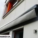 Markiza Moderno w kwadratowej kasecie o kolorze antracytowym z widoczną prostą falbaną w kolorze grafitowym zamocowana na ścianie budynku piętrowego o szarej nowoczesnej fasadzie z kontrastowymi pomarańczowymi elementami.