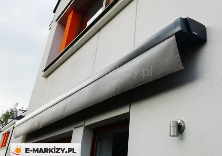 Markiza Moderno w kwadratowej kasecie o kolorze antracytowym z widoczną prostą falbaną w kolorze grafitowym zamocowana na ścianie budynku piętrowego o szarej nowoczesnej fasadzie z kontrastowymi pomarańczowymi elementami.