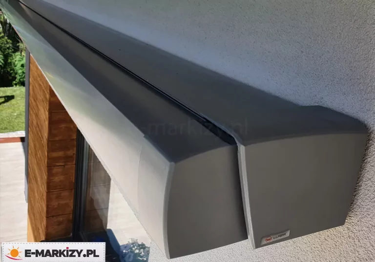 Widok z góry na kasetę oraz belkę przednią markizy Moderno MOL zamocowaną na ścianie nad oknem. Srebrna konstrukcja markizy kontrastuje z drewnianym wykończeniem w pobliżu okna.