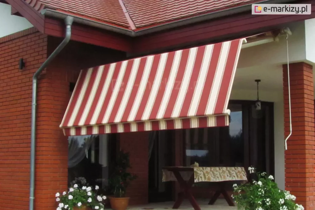 Zdjęcie prezentuje markizę tarasową Allegro zamontowaną we wnęce tarasowej domu jednorodzinnego o klasycznej architekturze z czerwonej cegły klinkierowej. Markiza jest w znacznym stopniu rozwinięta, a jej poszycie wykonane jest z estetycznej tkaniny w pionowe, regularne pasy w kolorach bordowym i kremowym. Cechą charakterystyczną tego modelu jest łamany profil ramion, który powoduje, że przednia część markizy opada niemal pionowo ku dołowi, skutecznie odcinając dopływ niskiego promieniowania słonecznego i osłaniając wnętrze tarasu oraz pomieszczeń. Konstrukcja wsporcza, obejmująca belkę montażową i ramiona, została polakierowana na neutralny biały kolor, który kontrastuje z ceglaną elewacją. Po prawej stronie widoczna jest biała korba służąca do ręcznego sterowania mechanizmem rozwijania. Markiza osłania strefę wypoczynkową, w której znajduje się drewniany stół przykryty wzorzystym obrusem. Wokół tarasu ustawione są donice z bujnie kwitnącymi białymi kwiatami, co w połączeniu z ciemnoczerwoną dachówką budynku tworzy spójną i przytulną aranżację przestrzeni zewnętrznej.