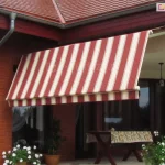 Zdjęcie prezentuje markizę tarasową Allegro zamontowaną we wnęce tarasowej domu jednorodzinnego o klasycznej architekturze z czerwonej cegły klinkierowej. Markiza jest w znacznym stopniu rozwinięta, a jej poszycie wykonane jest z estetycznej tkaniny w pionowe, regularne pasy w kolorach bordowym i kremowym. Cechą charakterystyczną tego modelu jest łamany profil ramion, który powoduje, że przednia część markizy opada niemal pionowo ku dołowi, skutecznie odcinając dopływ niskiego promieniowania słonecznego i osłaniając wnętrze tarasu oraz pomieszczeń. Konstrukcja wsporcza, obejmująca belkę montażową i ramiona, została polakierowana na neutralny biały kolor, który kontrastuje z ceglaną elewacją. Po prawej stronie widoczna jest biała korba służąca do ręcznego sterowania mechanizmem rozwijania. Markiza osłania strefę wypoczynkową, w której znajduje się drewniany stół przykryty wzorzystym obrusem. Wokół tarasu ustawione są donice z bujnie kwitnącymi białymi kwiatami, co w połączeniu z ciemnoczerwoną dachówką budynku tworzy spójną i przytulną aranżację przestrzeni zewnętrznej.