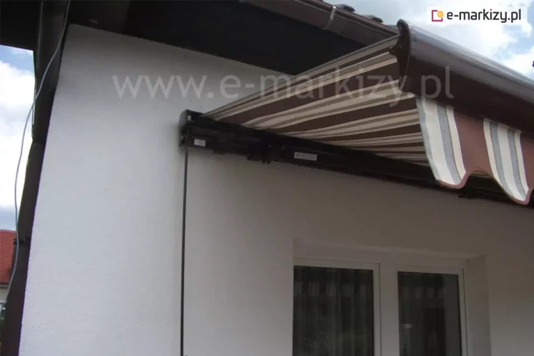 Zamocowana na ścianie o białym tynku, w pobliżu narożnika domu, tuz pod dachem, ale nad oknem, markiza tarasowa Palladio w kolorze brązowym z widoczną po jej lewej stronie korbą do sterowania ręcznego. Rozwinięte poszycie w brązowe, kremowe i szare pasy oraz falowana falbana zacieniają znajdujące się poniżej okno o białej ramie.