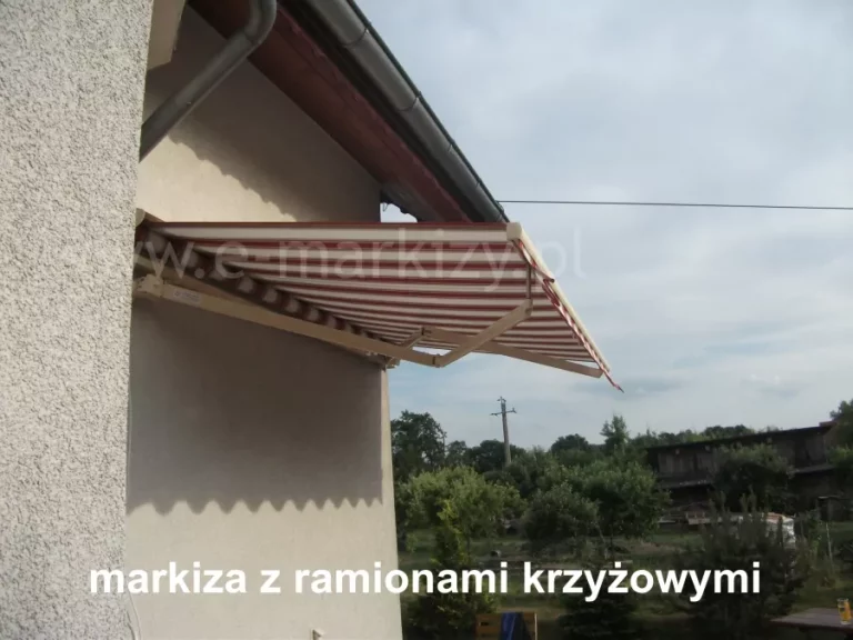 Zamocowana na jasnej ścianie domu, w pobliżu zakończenia dachu z rynną, markiza tarasowa Australia Aluprof bez kasety z aluminiową konstrukcją ramion krzyżowych w kolorze białym oraz poszyciem w biało-czerwone pasy. W tle za budynkiem widoczne krzewy i drzewa o zielonych liściach oraz słup elektryczny.