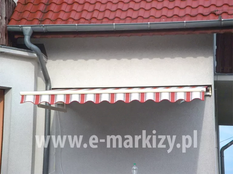 Markiza z widocznym po lewej stronie niższym ramieniem krzyżowym, zawieszona na ścianie z jasną elewacją pomiędzy narożnikiem budynku i rynną, nad markizą widoczny dach kryty czerwoną dachówką z szarą rynną