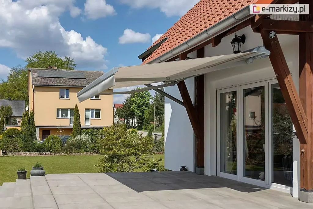 Zdjęcie prezentuje klasyczną markizę tarasową w pasy, zamontowaną na jasnej, tynkowanej elewacji piętrowego domu. Poszycie markizy charakteryzuje się naprzemiennymi, pionowymi pasami w kolorze białym i nasyconym żółtym, a jej krawędź wykończona jest ozdobną, pofalowaną falbaną o tym samym wzorze. Konstrukcja markizy oraz listwa frontowa są w kolorze białym, co koresponduje z ramami dużych, przeszklonych drzwi tarasowych i okien z podziałami szprosowymi. Markiza jest w pełni rozłożona, zacieniając przestronny taras wyłożony dużymi płytami kamiennymi w odcieniach beżu i szarości. W bocznej części tarasu znajduje się rozbudowany, murowany grill zewnętrzny lub wędzarnia z cegły klinkierowej, a w tle rozciąga się widok na zielone pastwiska i pagórkowaty krajobraz pod zachmurzonym niebem. Na pierwszym planie widoczne są dwa małe psy oraz donice z roślinnością, co podkreśla domowy i rekreacyjny charakter prezentowanej przestrzeni.