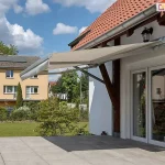 Zdjęcie prezentuje klasyczną markizę tarasową w pasy, zamontowaną na jasnej, tynkowanej elewacji piętrowego domu. Poszycie markizy charakteryzuje się naprzemiennymi, pionowymi pasami w kolorze białym i nasyconym żółtym, a jej krawędź wykończona jest ozdobną, pofalowaną falbaną o tym samym wzorze. Konstrukcja markizy oraz listwa frontowa są w kolorze białym, co koresponduje z ramami dużych, przeszklonych drzwi tarasowych i okien z podziałami szprosowymi. Markiza jest w pełni rozłożona, zacieniając przestronny taras wyłożony dużymi płytami kamiennymi w odcieniach beżu i szarości. W bocznej części tarasu znajduje się rozbudowany, murowany grill zewnętrzny lub wędzarnia z cegły klinkierowej, a w tle rozciąga się widok na zielone pastwiska i pagórkowaty krajobraz pod zachmurzonym niebem. Na pierwszym planie widoczne są dwa małe psy oraz donice z roślinnością, co podkreśla domowy i rekreacyjny charakter prezentowanej przestrzeni.