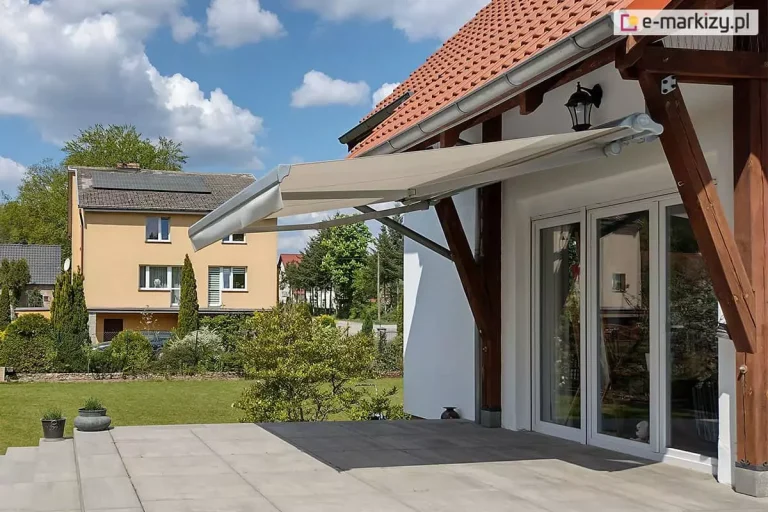 Zdjęcie prezentuje klasyczną markizę tarasową w pasy, zamontowaną na jasnej, tynkowanej elewacji piętrowego domu. Poszycie markizy charakteryzuje się naprzemiennymi, pionowymi pasami w kolorze białym i nasyconym żółtym, a jej krawędź wykończona jest ozdobną, pofalowaną falbaną o tym samym wzorze. Konstrukcja markizy oraz listwa frontowa są w kolorze białym, co koresponduje z ramami dużych, przeszklonych drzwi tarasowych i okien z podziałami szprosowymi. Markiza jest w pełni rozłożona, zacieniając przestronny taras wyłożony dużymi płytami kamiennymi w odcieniach beżu i szarości. W bocznej części tarasu znajduje się rozbudowany, murowany grill zewnętrzny lub wędzarnia z cegły klinkierowej, a w tle rozciąga się widok na zielone pastwiska i pagórkowaty krajobraz pod zachmurzonym niebem. Na pierwszym planie widoczne są dwa małe psy oraz donice z roślinnością, co podkreśla domowy i rekreacyjny charakter prezentowanej przestrzeni.