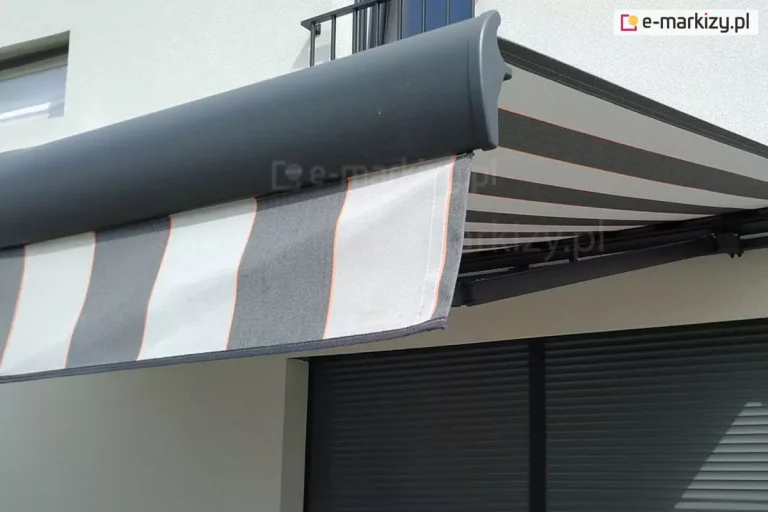 Markiza Palladio Aluprof w kolorze grafitowym z tkaniną w szare i jasnoszare pasy z cienkimi liniami w kolorze czerwonym zamocowana została na białej ścianie domu nad dużym oknem tarasowym przesłoniętym roletą zewnętrzną. Rozwinięte poszycie rzuca cień na ścianę i okno.