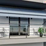 Zdjęcie przedstawia nowoczesny balkon wyposażony w rozłożystą markizę balkonowo-sklepową oraz balustradę ze stali nierdzewnej. Markiza w kolorze jasnoszarym jest zamontowana do białej elewacji budynku pod nawisem kolejnej kondygnacji. Materiał poszycia jest gładki i podzielony na cztery pionowe segmenty, a jej konstrukcja opiera się na bocznych ramionach, które stabilizują wysięg nad przestrzenią wypoczynkową. Poniżej markizy znajduje się okno balkonowe o ciemnych ramach, w którym odbija się otoczenie. Balustrada wykonana z błyszczącego metalu składa się z poziomych prętów zamocowanych do pionowych słupków, tworząc ażurową osłonę balkonu. Na podłodze balkonu, która wyłożona jest jasnymi płytkami, ustawiono trzy dekoracyjne rośliny w ciemnych donicach o zaokrąglonym kształcie. Rośliny mają gęste, zielone liście i są rozmieszczone symetrycznie – jedna po lewej stronie oraz dwie po prawej. W głębi, za balustradą, widoczne są dwa jasne krzesła ogrodowe o plecionej strukturze oparć. Całość aranżacji utrzymana jest w minimalistycznym i nowoczesnym stylu, z dominacją bieli, szarości i metalicznych akcentów.