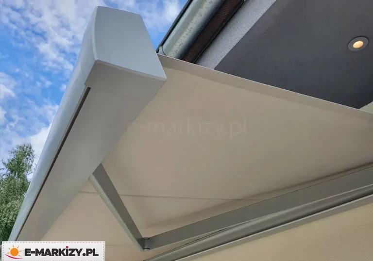 Markiza w kwadratowej kasecie bez falbany model Moderno mol ze srebrną konstrukcją oraz kremowym poszyciem rozwinięta pod okapem dachu w szarym kolorze z widocznym punktem świetlnym. Ponad markizą widoczne niebieskie niebo lekko pokryte białymi chmurkami, a obok belki przedniej z lewej strony drzewo o zielonych liściach.