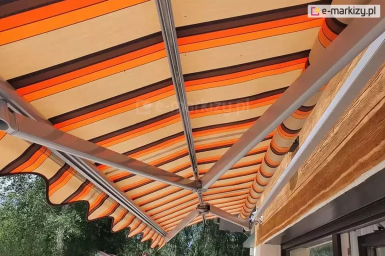 Zdjęcie prezentuje techniczne zbliżenie na markizę tarasową Allegro, skupiając się na detalach wykończenia profilu czołowego, belce środkowej oraz strukturze tkaniny. Widoczna jest solidna belka przednia wykonana z aluminium lakierowanego na kolor jasnoszary, która domyka konstrukcję i służy do stabilnego naciągu poszycia. Belka środkowa odpowiada za odpowiednia prace ramion, zapewniając unikalny łamany wysięg markizy Allegro. Materiał markizy charakteryzuje się wzorem w pionowe, kontrastujące pasy o nasyconych barwach pomarańczowego, ciemnego brązu oraz piaskowego beżu. Tkanina wykazuje wyraźny, gęsty splot, co świadczy o jej wysokiej odporności na warunki atmosferyczne i promieniowanie UV. W prawym dolnym rogu kadru, poza obrysem osłony, widoczne są rozmyte korony drzew liściastych na tle jasnego, lekko zachmurzonego nieba, co podkreśla funkcjonalność produktu w przestrzeni ogrodowej. Fotografia kładzie nacisk na estetykę połączenia metalowych elementów konstrukcyjnych z tekstylnym wykończeniem osłony.