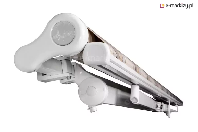 Zdjęcie prezentuje na białym tle produkt, którym jest markiza Mezzo w pozycji złożonej, ukazaną z perspektywy bocznej pod kątem. Aluminiowy stelaż w kolorze białym składa się z dekoracyjnej bocznej pokrywy rury nawojowej z subtelnym wzorem graficznym, belki frontowej oraz złożonych ramion przegubowych. Pomiędzy profilami widoczny jest zwinięty materiał poszycia w pasy o beżowej kolorystyce. Konstrukcja wsparta jest na solidnym uchwycie montażowym z widocznymi śrubami regulacyjnymi, co podkreśla stabilność i wysoką jakość wykonania mechanizmu. Całość tworzy zwartą i estetyczną formę, która po złożeniu zajmuje minimalną ilość miejsca na elewacji.
