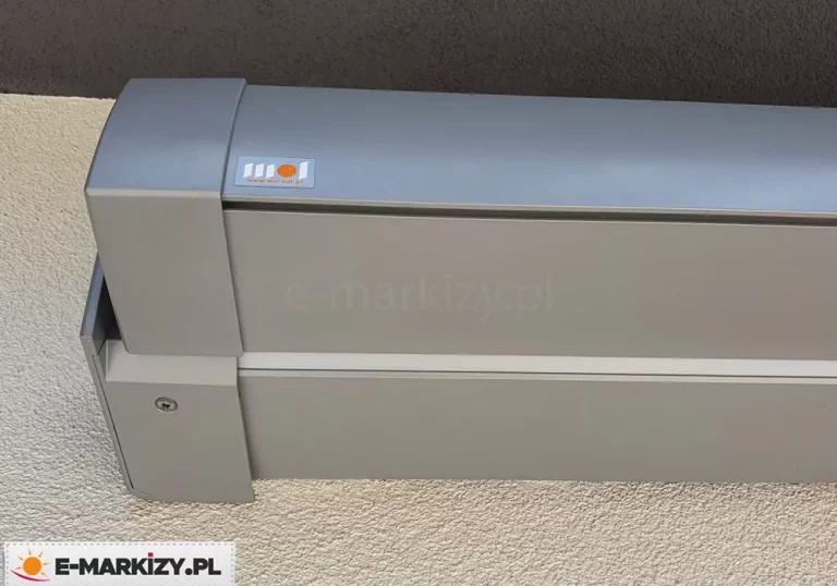 Markizy firmy MOL model Moderno z widocznym logo producenta na skraju belki przedniej po lewej stronie. Charakterystyczna kwadratowa kaseta w srebrnym kolorze zamocowana została do ściany pokrytej białym tynkiem strukturalnym.