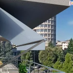 Zdjęcie prezentuje zbliżenie od boku na konstrukcję markizy balkonowej zamontowanej na jasnej elewacji budynku wielorodzinnego. Markiza o jasnoszarym poszyciu jest częściowo wysunięta, chroniąc przestrzeń balkonu przed słońcem. Widoczne są solidne, ciemnoszare ramiona boczne mocowane bezpośrednio do ściany za pomocą metalowych uchwytów, które stabilizują materiał. Górna część mechanizmu znajduje się pod krawędzią wyższej kondygnacji, co zapewnia dodatkową ochronę dla samej kasety urządzenia. Na balkonie widoczna jest nowoczesna balustrada z połyskujących, poziomych profili metalowych, za którą znajduje się fragment plecionego fotela oraz donica z gęstą, zieloną roślinnością. W tle rozpościera się widok na miejską panoramę, w tym na charakterystyczny wysoki budynek o cylindrycznym kształcie z licznymi oknami, otoczony przez korony drzew i niższe bloki mieszkalne. Perspektywa zdjęcia została ujęta z boku, co pozwala dokładnie przyjrzeć się kątowi nachylenia materiału markizy oraz elementom montażowym przytwierdzonym do białego tynku budynku.