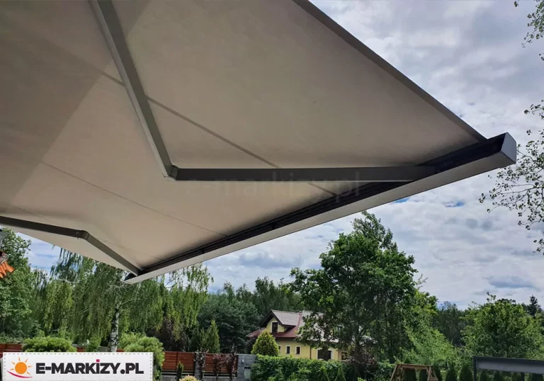 Zdjęcie przedstawia nowoczesną markizę tarasową Moderno w pełnej kasecie, widoczną od strony dolnej mechanizmu oraz poszycia. Konstrukcja opiera się na dwóch solidnych, przegubowych ramionach bocznych wykonanych z metalu o matowym wykończeniu w kolorze ciemnoszarym lub antracytowym. Ramiona te podtrzymują rozciągniętą tkaninę poszycia w kolorze jasnokremowym lub piaskowym, która jest napięta i tworzy jednolitą płaszczyznę osłaniającą przestrzeń przed słońcem. Na krawędzi zewnętrznej widoczna jest estetyczna, prostokątna listwa przednia o ostrych, nowoczesnych liniach, która po złożeniu markizy szczelnie domyka kasetę. W tle znajduje się ogród z gęstą roślinnością, drzewami liściastymi oraz fragmentem domu jednorodzinnego o żółtej elewacji i ciemnym dachu, a nad całością rozciąga się lekko zachmurzone niebo.