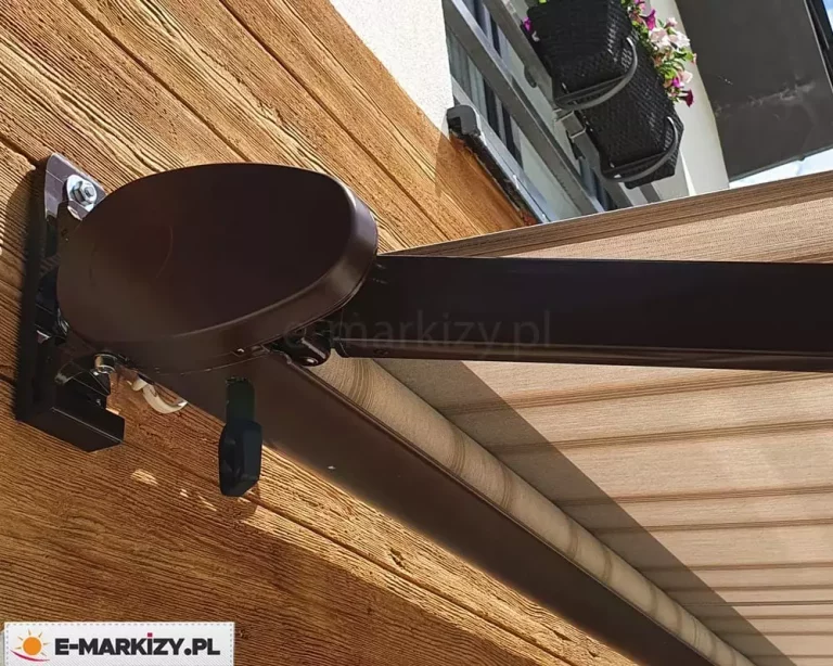 Zdjęcie prezentuje zbliżenie na techniczne detale montażowe oraz mechanizm boczny rozłożonej markizy tarasowej. Kluczowym elementem jest masywny, owalny uchwyt boczny w kolorze ciemnobrązowym, który łączy ramię przegubowe z kasetą markizy. Tuż pod kasetą widoczny jest czarny uchwyt przekładni korbowej do napędu ręcznego. Mechanizm jest solidnie przytwierdzony do elewacji budynku za pomocą stalowych śrub widocznych w górnej części wspornika. Ściana, na której zamontowano urządzenie, wyłożona jest dekoracyjnymi panelami o wyraźnej strukturze drewna w ciepłym, miodowym odcieniu. Z mechanizmu wysuwa się ramię o profilu prostokątnym, podtrzymujące napięte poszycie wykonane z tkaniny w kolorze jasnego beżu z delikatnymi, poziomymi prążkami. Poniżej głównego profilu widoczna jest rura nawojowa z nawiniętym materiałem. W tle, w górnej części kadru, dostrzec można fragment białej elewacji wyższej kondygnacji oraz czarną skrzynkę balkonową wypełnioną kwitnącymi kwiatami w kolorze białym i fioletowym. Perspektywa zdjęcia z bliska pozwala dokładnie zaobserwować jakość wykończenia powłoki lakierniczej stelaża oraz precyzję dopasowania poszczególnych komponentów mechanicznych markizy.