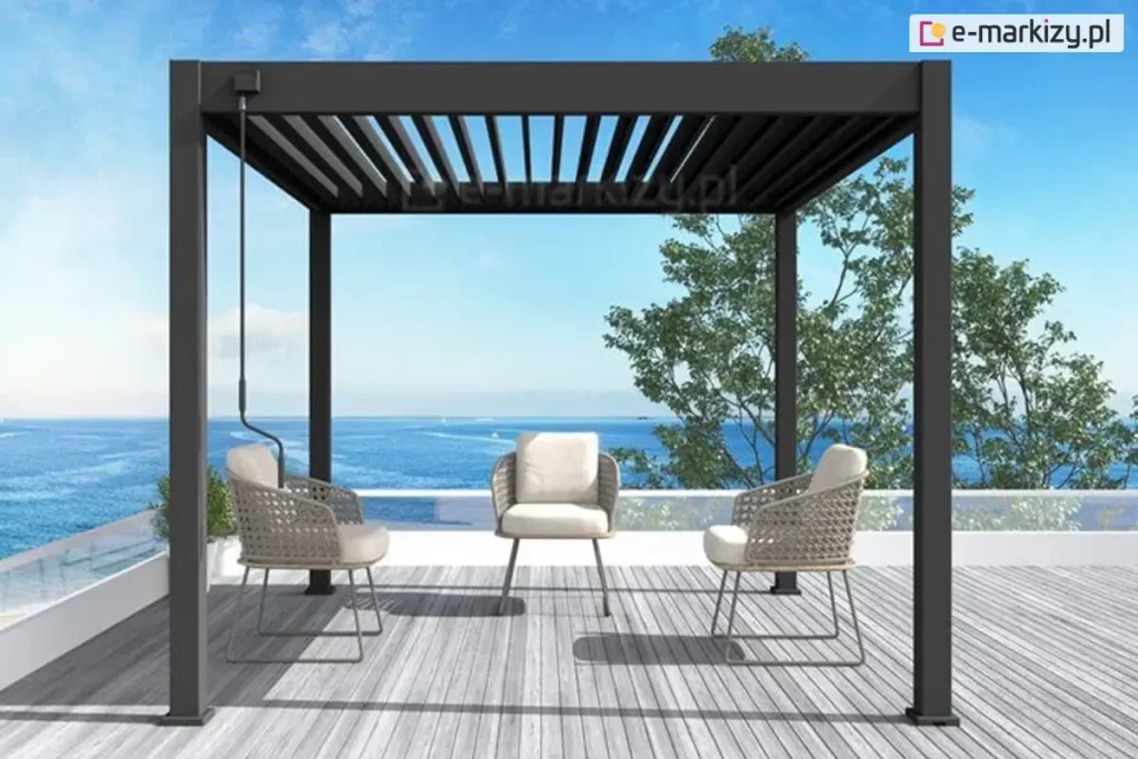 Wolnostojąca pergola lamelowa Marlo w kolorze antracytowym, zainstalowana na nowoczesnym tarasie z widokiem na morze. Konstrukcja o kwadratowym przekroju słupów posiada dach wykonany z ruchomych lameli aluminiowych, które są ustawione w pozycji częściowo otwartej. Taras wyłożony jest deskami kompozytowymi w kolorze jasnoszarym i zabezpieczony szklaną balustradą z białą ramą, co zapewnia nieograniczony widok na błękitną wodę oraz horyzont. Pod zadaszeniem znajduje się minimalistyczna strefa wypoczynkowa składająca się z trzech foteli o ażurowym, beżowym splocie z białymi poduszkami siedziskowymi. Przy lewym frontowym słupku konstrukcyjnym zamontowana jest czarna korba służąca do ręcznej regulacji kąta nachylenia dachu. W tle, po prawej stronie, widoczne są zielone korony drzew liściastych na tle czystego, błękitnego nieba.