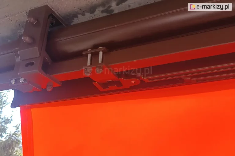 Szczegółowe zbliżenie na techniczne aspekty montażu markizy tarasowej do powierzchni sufitu, widziane od spodu konstrukcji. Głównym elementem kadru jest aluminiowy uchwyt montażowy w kolorze brązowym, który mocuje profil nośny markizy do betonowego stropu lub nadproża o surowej, porowatej strukturze. Uchwyt jest solidnie przytwierdzony za pomocą dwóch śrub z ozdobnymi, półkolistymi nakrętkami typu kołpakowego do części uchwytu ściennego, w który wsunięta jest belka kwadratowa (nośna) markizy. Widoczne są również inne elementy mechanizmu, w tym mocowanie ramion oraz krawędź kasety. Z lewej strony kadru znajduje się fragment rozwiniętego poszycia markizy w intensywnym, żółto-pomarańczowym kolorze, które kontrastuje z metalicznym wykończeniem osprzętu i szarością betonu. Zdjęcie ma charakter techniczny i dokumentacyjny, pokazując precyzję wykonania instalacji oraz sposób stabilizacji całego systemu osłonowego. Perspektywa z dołu pozwala na dokładną ocenę odstępu konstrukcji od ściany oraz jakości użytych komponentów złącznych.
