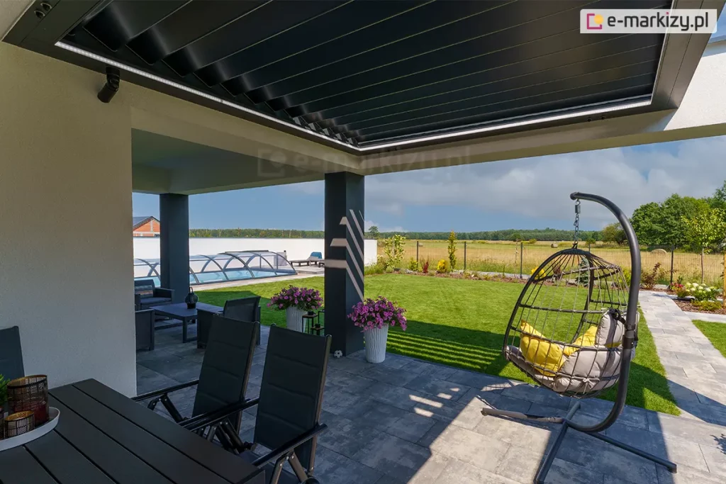 Zdjęcie prezentuje perspektywę z wnętrza tarasu skierowaną na ogród, ukazującą bioklimatyczny dach lamelowy SB400R w pozycji lekko uchylonej. Aluminiowe lamele w kolorze antracytowym tworzą jednolitą płaszczyznę zadaszenia, chroniącą przed słońcem i deszczem. Wzdłuż wewnętrznej krawędzi ramy konstrukcyjnej zamontowano liniowe oświetlenie LED, które emituje jasne, białe światło, doświetlając przestrzeń pod pergolą. Konstrukcja wsparta jest na masywnym, ciemnoszarym słupie nośnym, a posadzka tarasu wykonana jest z dużych, jasnoszarych płyt betonowych. W strefie wypoczynkowej widoczny jest wiszący fotel ogrodowy typu kokon z szarymi poduszkami oraz czarne krzesła przy stole jadalnianym. W tle rozpościera się widok na zadbany ogród z zielonym trawnikiem, basenem przykrytym niską, transparentną kopułą oraz otwartą przestrzeń pól pod błękitnym niebem z rozproszonymi chmurami.