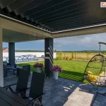 Zdjęcie prezentuje perspektywę z wnętrza tarasu skierowaną na ogród, ukazującą bioklimatyczny dach lamelowy SB400R w pozycji lekko uchylonej. Aluminiowe lamele w kolorze antracytowym tworzą jednolitą płaszczyznę zadaszenia, chroniącą przed słońcem i deszczem. Wzdłuż wewnętrznej krawędzi ramy konstrukcyjnej zamontowano liniowe oświetlenie LED, które emituje jasne, białe światło, doświetlając przestrzeń pod pergolą. Konstrukcja wsparta jest na masywnym, ciemnoszarym słupie nośnym, a posadzka tarasu wykonana jest z dużych, jasnoszarych płyt betonowych. W strefie wypoczynkowej widoczny jest wiszący fotel ogrodowy typu kokon z szarymi poduszkami oraz czarne krzesła przy stole jadalnianym. W tle rozpościera się widok na zadbany ogród z zielonym trawnikiem, basenem przykrytym niską, transparentną kopułą oraz otwartą przestrzeń pól pod błękitnym niebem z rozproszonymi chmurami.
