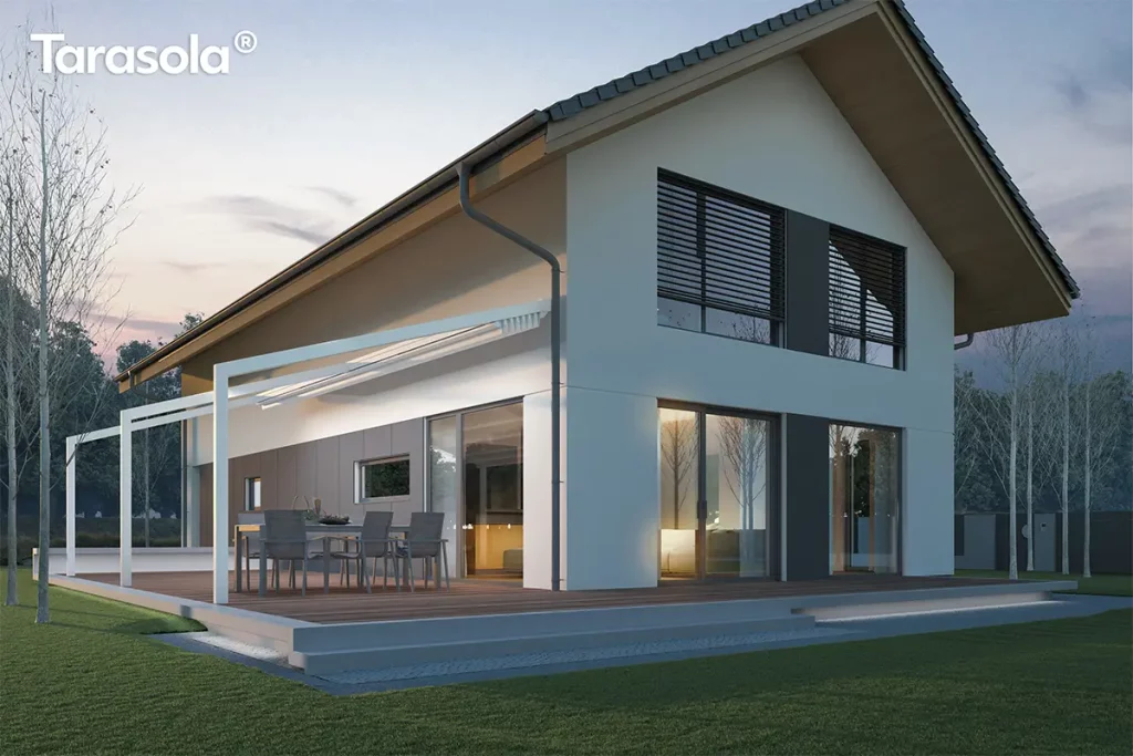 Nowoczesna pergola tarasowa Tarasola Light w kolorze białym zamontowana do elewacji piętrowego domu jednorodzinnego w stylu stodoły. Konstrukcja posiada zadaszenie z jasnej tkaniny technicznej, które jest częściowo zwinięte, ukazując lekkość aluminiowego stelaża wspartego na smukłych nogach o kwadratowym przekroju. Pod zadaszeniem znajduje się strefa jadalniana z szarym stołem i krzesłami ogrodowymi, ustawiona na drewnianym podestowym tarasie zintegrowanym z bryłą budynku. Budynek posiada białą elewację z antracytowymi akcentami przy oknach oraz duże przeszklenia, przez które przebija ciepłe, domowe światło. Wieczorna sceneria podkreśla subtelne oświetlenie LED zamontowane w profilach tkaniny oraz liniowe podświetlenie schodów prowadzących na taras. Wokół domu rozciąga się równo przystrzyżony trawnik oraz szpaler młodych drzew na tle przygasającego, wieczornego nieba.