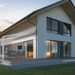 Nowoczesna pergola tarasowa Tarasola Light w kolorze białym zamontowana do elewacji piętrowego domu jednorodzinnego w stylu stodoły. Konstrukcja posiada zadaszenie z jasnej tkaniny technicznej, które jest częściowo zwinięte, ukazując lekkość aluminiowego stelaża wspartego na smukłych nogach o kwadratowym przekroju. Pod zadaszeniem znajduje się strefa jadalniana z szarym stołem i krzesłami ogrodowymi, ustawiona na drewnianym podestowym tarasie zintegrowanym z bryłą budynku. Budynek posiada białą elewację z antracytowymi akcentami przy oknach oraz duże przeszklenia, przez które przebija ciepłe, domowe światło. Wieczorna sceneria podkreśla subtelne oświetlenie LED zamontowane w profilach tkaniny oraz liniowe podświetlenie schodów prowadzących na taras. Wokół domu rozciąga się równo przystrzyżony trawnik oraz szpaler młodych drzew na tle przygasającego, wieczornego nieba.